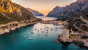 Vue aérienne réaliste de la marine de Porto en Corse, avec une eau turquoise, un golfe entouré de montagnes et une tour génoise au coucher du soleil.